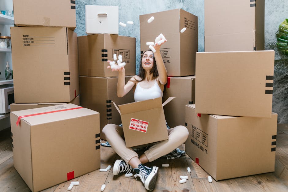 A young woman with long brown hair, dressed in a white tank top and beige trousers, is sitting cross-legged on a wooden floor inside a home, surrounded by numerous large cardboard moving boxes. She is smiling and playfully tossing packing foam peanuts into the air, indicating a packing or unpacking process as part of a home relocation. The boxes vary in size, some with red tape and labels, and are stacked against a light-colored wall with textured blue and grey paint. A few boxes are positioned on the floor, while others are stacked on top of each other. In the background, there are household items, including a small white refrigerator and a potted plant, and an open door leading into another room. The scene is well-lit with natural light, reinforcing a context of furniture transport and packing within a moving and removals service environment, such as Pimlico Removals, involved in house moves and logistics.