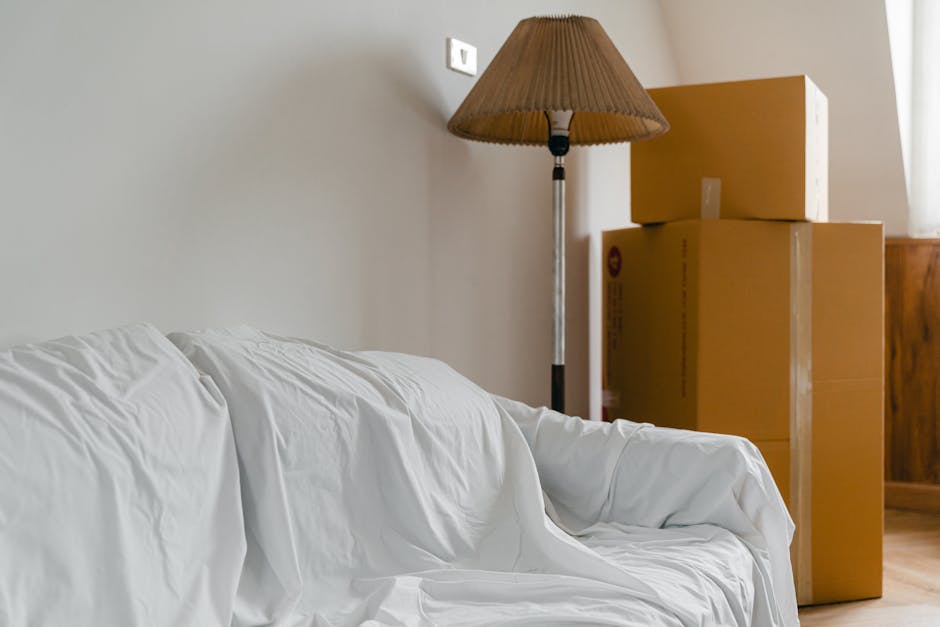 A living room interior showing a white sofa draped with a white blanket on the left side of the image, with three cardboard moving boxes stacked behind it near the right wall. The boxes are sealed with brown packing tape, with the smallest on top, the medium-sized box beneath it, and a larger box at the bottom. A tall, standing floor lamp with a beige shade is positioned between the sofa and the boxes, providing ambient lighting to the space. The background features a plain light-colored wall with a power socket visible above the boxes, and a window with sheer curtains letting natural light into the room. This scene illustrates part of a home relocation process, with packing supplies and furniture ready for moving services often offered by companies like Pimlico Removals, as indicated by the context of the webpage about bulky furniture removals in Pimlico.