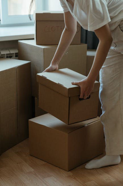 A young woman with long brown hair, dressed in a white tank top and beige trousers, is sitting cross-legged on a wooden floor inside a home, surrounded by numerous large cardboard moving boxes. She is smiling and playfully tossing packing foam peanuts into the air, indicating a packing or unpacking process as part of a home relocation. The boxes vary in size, some with red tape and labels, and are stacked against a light-colored wall with textured blue and grey paint. A few boxes are positioned on the floor, while others are stacked on top of each other. In the background, there are household items, including a small white refrigerator and a potted plant, and an open door leading into another room. The scene is well-lit with natural light, reinforcing a context of furniture transport and packing within a moving and removals service environment, such as Pimlico Removals, involved in house moves and logistics.
