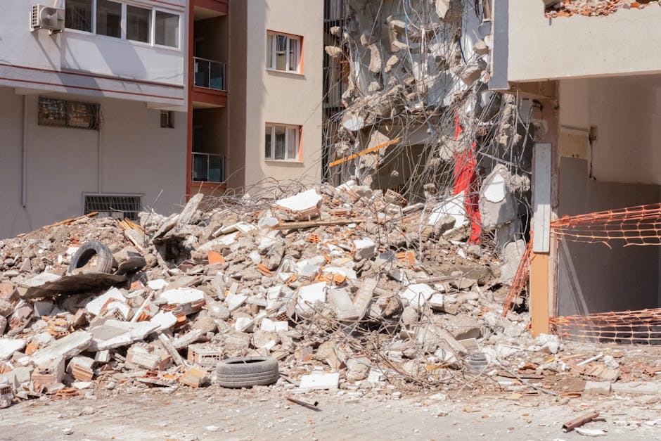 An exterior scene showing a large pile of demolition debris consisting of broken concrete, bricks, twisted metal rods, and scattered construction materials accumulated at the base of a partially demolished residential building. The debris, including a black tire and pieces of wooden planks, is situated on a dirt ground next to the building's entrance. The surrounding area features an orange safety fence and construction mesh, with the building's facade displaying exposed structural elements and some intact windows. The image illustrates the aftermath of renovation or demolition work, which is relevant to the context of house removals and clearing waste after renovation projects, as handled by Pimlico Removals.
