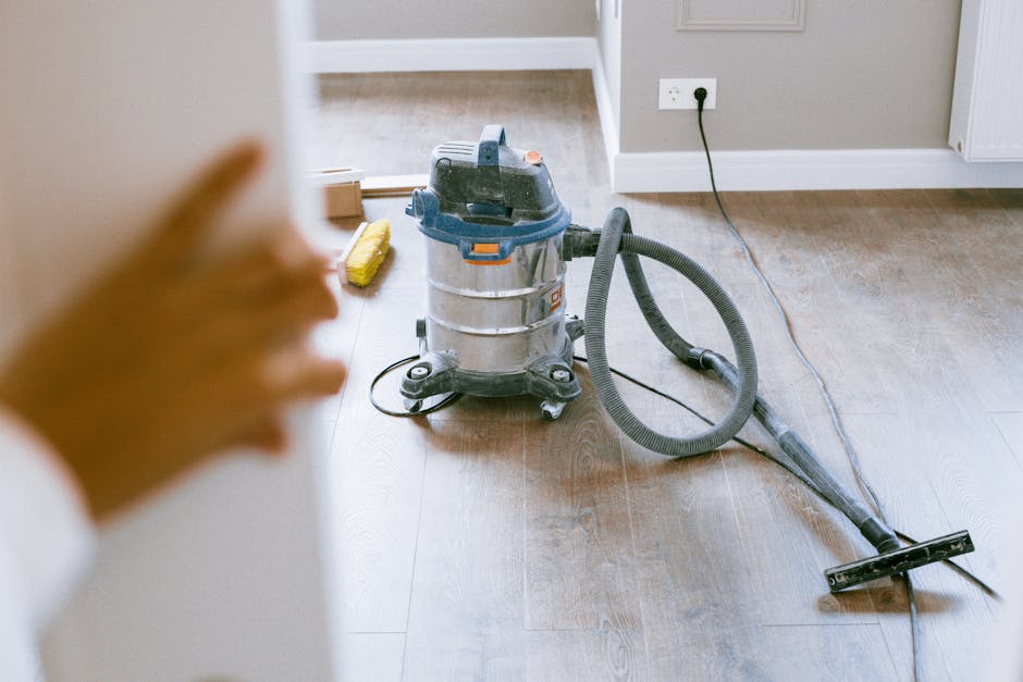 A close-up view of a professional vacuum cleaner with a metallic silver body, positioned on a wooden floor inside a residential property. The vacuum has a blue top and a flexible hose attached to a nozzle, which is resting on the floor. Nearby, a yellow scrubbing brush and a cardboard box are partially visible in the background. The vacuum cleaner is plugged into a wall socket, with the cord extending across the floor. The scene depicts a cleaning process relevant to house cleaning or waste removal during home relocation or renovation activities, with natural ambient lighting illuminating the workspace. The setting reflects an indoor environment where cleaning materials and equipment are prepared for waste clearance, supporting services such as packing and moving by Pimlico Removals.