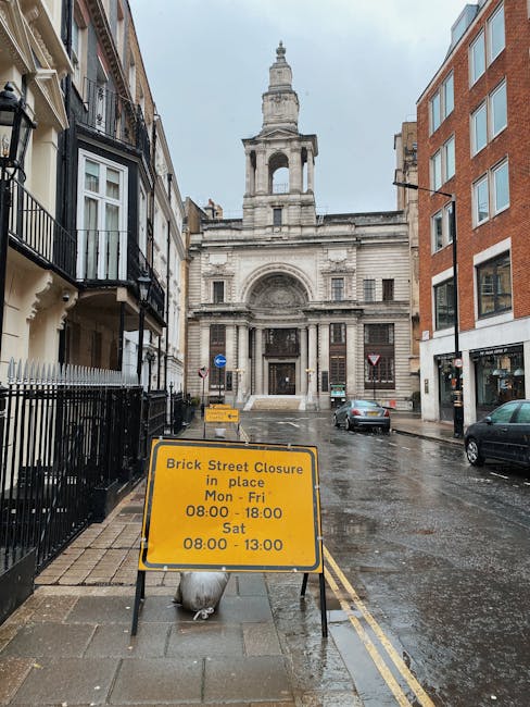 A narrow street scene in an urban area during rainy weather, with wet pavement and reflections visible on the ground. On the left side, there are traditional Victorian-style terraced houses with black railings and small balconies. On the right, modern brick buildings with large windows line the street. In the background, a historic stone church with a prominent tower and classical architectural details stands at the end of the street. A yellow road closure sign is placed on the sidewalk in the foreground, indicating street closure times from Monday to Saturday, with the mention of a brick street closure in place. Vehicle traffic includes a few parked cars and a moving dark grey car. The overall scene depicts a typical street in central London, suitable for home relocation or furniture transport activities, with Pimlico Removals providing professional removals services in the area.