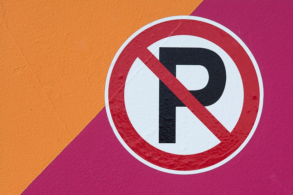 Close-up of a round, red-bordered 'No Parking' sign with a bold black 'P' crossed out in red, mounted on a vibrant wall painted with large patches of orange, pink, and purple. The sign is positioned on the corner of the wall near a doorway or pavement area, indicating parking restrictions relevant to house removals and moving services performed by Pimlico Removals. The surface of the wall shows a textured finish with small cracks, and the lighting is even, highlighting the sign and the colorful background, which emphasizes the importance of parking regulations during the home relocation or furniture transport process managed by the company.