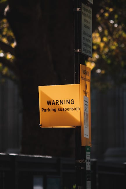 A yellow warning sign attached to a metal pole displays the message 'WARNING Parking suspension' in black text. The sign is illuminated by natural daylight, with a blurred background featuring trees, foliage, and a metal fence. The pole also holds other notices and parking-related information. This signage indicates that parking restrictions are in effect in the area, which is relevant for house removals and moving services provided by Pimlico Removals. The image captures the urban environment where temporary parking suspensions are common during home relocation or furniture transport activities, emphasizing the importance of checking parking regulations before moving. The environment appears quiet, with soft sunlight filtering through the leaves, and the focus remains on the clear, visible warning sign necessary for logistics planning in house removals operations.