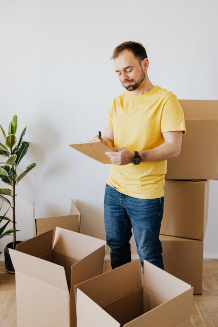 A man with short dark hair and a beard, wearing a yellow t-shirt and blue jeans, is standing inside a room preparing for a home relocation. He is holding a clipboard and a pen, inspecting or making notes about the packing process. Surrounding him are several open cardboard boxes, some on the floor and others stacked against the wall, ready for loading. The room has a white wall, a tall green plant in a pot on the left, and a wooden floor. Light from an unseen source illuminates the scene evenly, highlighting the organized packing materials. Large cardboard boxes, typical for furniture transport and moving logistics, are being prepared for loading or unpacking, reflecting a professional approach to furniture transport and packing during an urgent move. This image illustrates the packing and moving process that Pimlico Removals offers as part of their removals services.