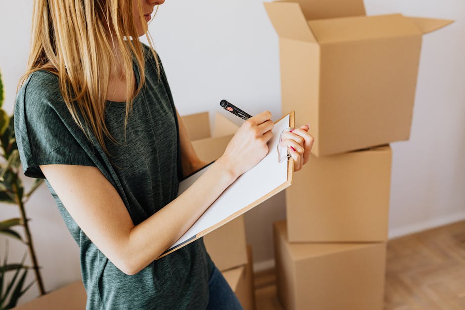 A woman with long blonde hair wearing a dark green T-shirt is inside a room, writing on a clipboard with a black marker. She is positioned next to several large, sealed cardboard boxes stacked on top of each other on a wooden floor. The background features a plain white wall and a potted plant with green leaves. The boxes are unlabelled and appear to be ready for a house move, as part of a home relocation or packing process. The room is well-lit with natural or soft artificial light, and the scene captures the logistics involved in packing and preparing belongings for furniture transport or an urgent move, consistent with services offered by Pimlico Removals in the context of same-day removals.