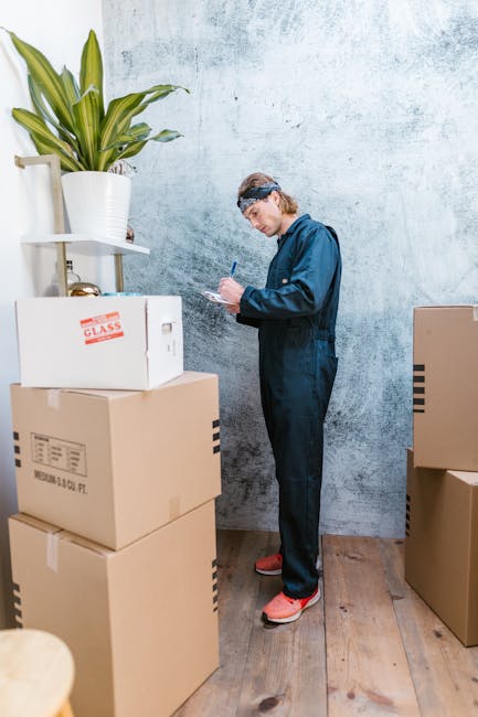 A man with short dark hair and a beard, wearing a yellow t-shirt and blue jeans, is standing inside a room preparing for a home relocation. He is holding a clipboard and a pen, inspecting or making notes about the packing process. Surrounding him are several open cardboard boxes, some on the floor and others stacked against the wall, ready for loading. The room has a white wall, a tall green plant in a pot on the left, and a wooden floor. Light from an unseen source illuminates the scene evenly, highlighting the organized packing materials. Large cardboard boxes, typical for furniture transport and moving logistics, are being prepared for loading or unpacking, reflecting a professional approach to furniture transport and packing during an urgent move. This image illustrates the packing and moving process that Pimlico Removals offers as part of their removals services.