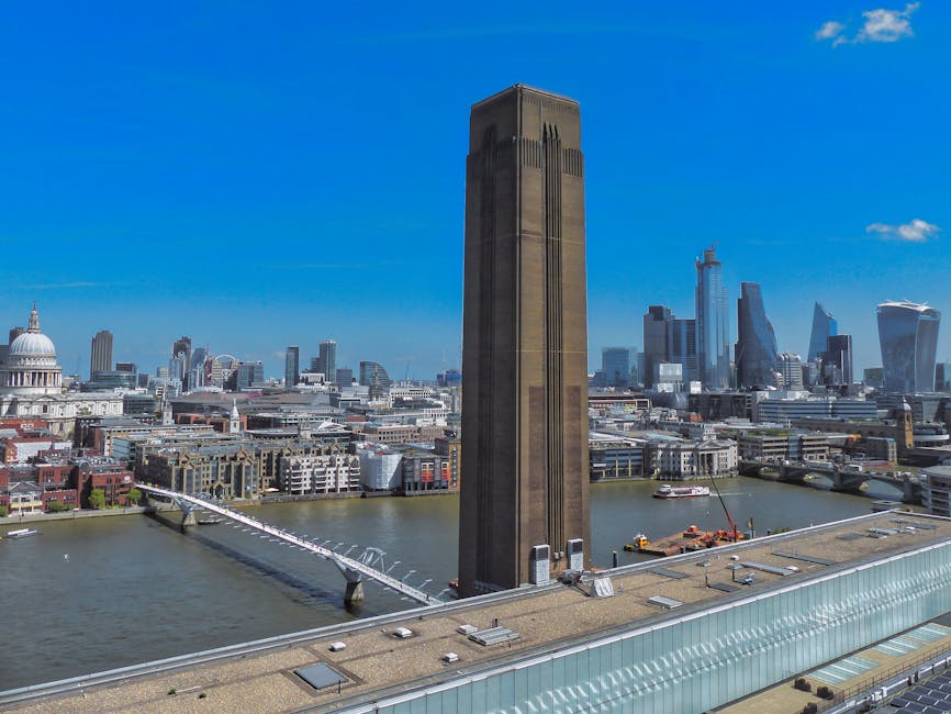 A high-angle view of the London skyline featuring The Leadenhall Building, also known as the Cheesegrater, with a clear blue sky in the background. The River Thames is visible at the bottom of the image, with a bridge crossing over it. In the foreground, a flat roof area with small ventilation units and rooftop equipment can be seen, indicating the vantage point is from a tall building. The cityscape includes various modern skyscrapers, historic structures, and office buildings. The scene captures the urban environment typical of central London, relevant to house removals and moving logistics in the city. Pimlico Removals often facilitates furniture transport and home relocation services near iconic landmarks like these, ensuring smooth packing and loading processes during moving projects like the Tate Britain to Riverside moves.