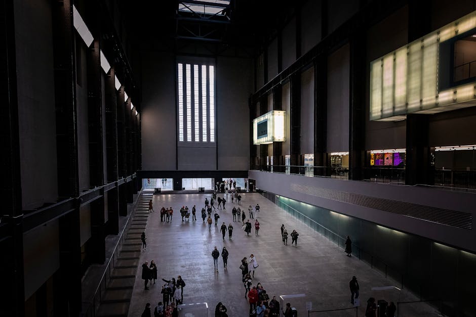 The image depicts the interior of a spacious, modern gallery or museum with high ceilings and large vertical windows allowing natural light to filter in. The polished, light-colored floor is occupied by numerous visitors walking and observing the space, some in groups and others alone. Along the left side, there are tall, dark structural columns and a series of evenly spaced display panels or artwork. On the right, a glass-paneled balcony runs along the upper level, overlooking the main hall, with digital screens mounted on the upper wall. The background shows a large, high window and an entrance area at the far end, where a few visitors are entering or leaving. The scene is well-lit, with a combination of natural and artificial lighting. occasional cardboard boxes, packing materials, and small furniture pieces are visible on the ground, indicating the gallery may be in the process of moving or setting up. Recognisable as part of professional moving logistics, possibly related to a house relocation or gallery packing and moving. The image reflects a loading process or moving activity, consistent with a professional removals service such as Pimlico Removals, supporting efficient furniture transport and packing within a public or exhibition space.